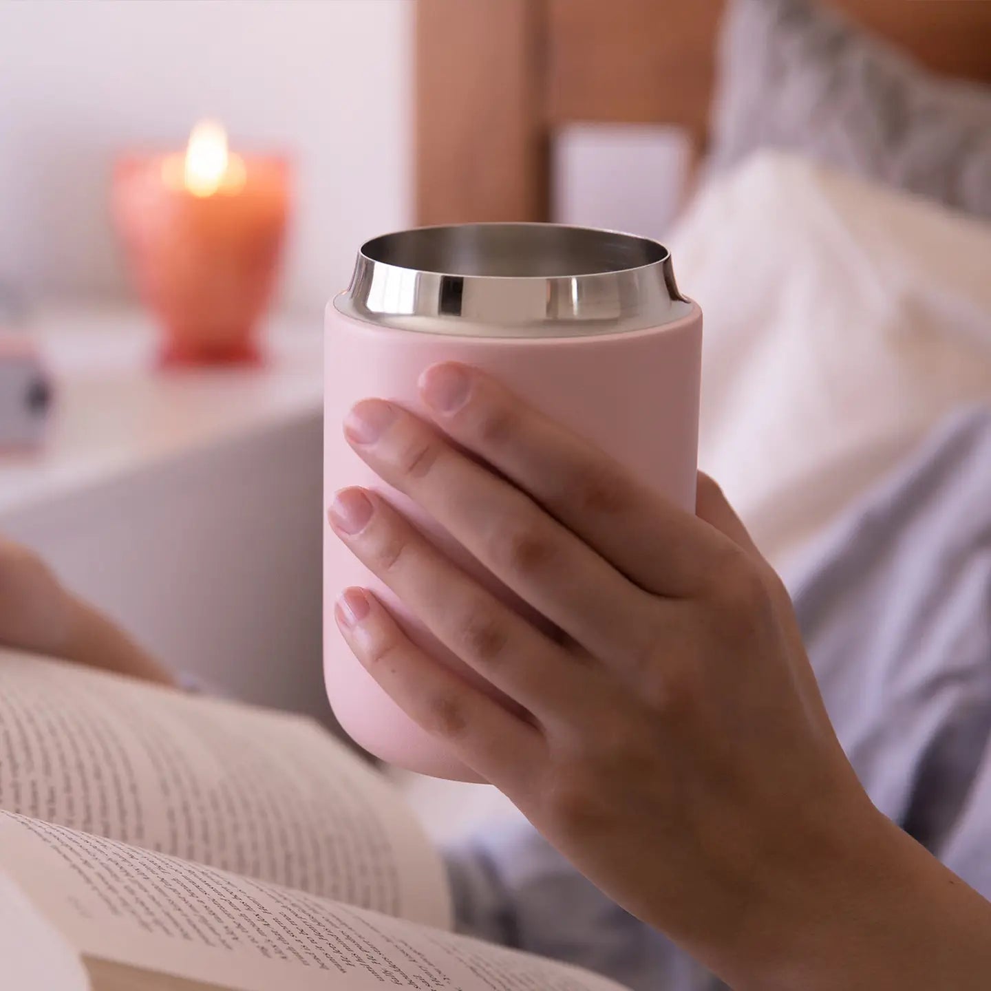 A matte pink, wide-mouth travel mug with a polished stainless steel rim, held in a person's hand over an open book.