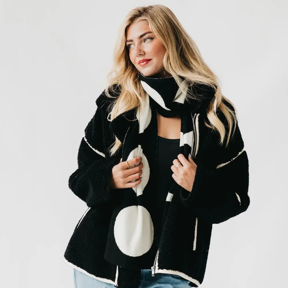 A woman in a black fuzzy sherpa jacket wears a thick black and white oversized polka dot scarf draped loosely around her neck.
