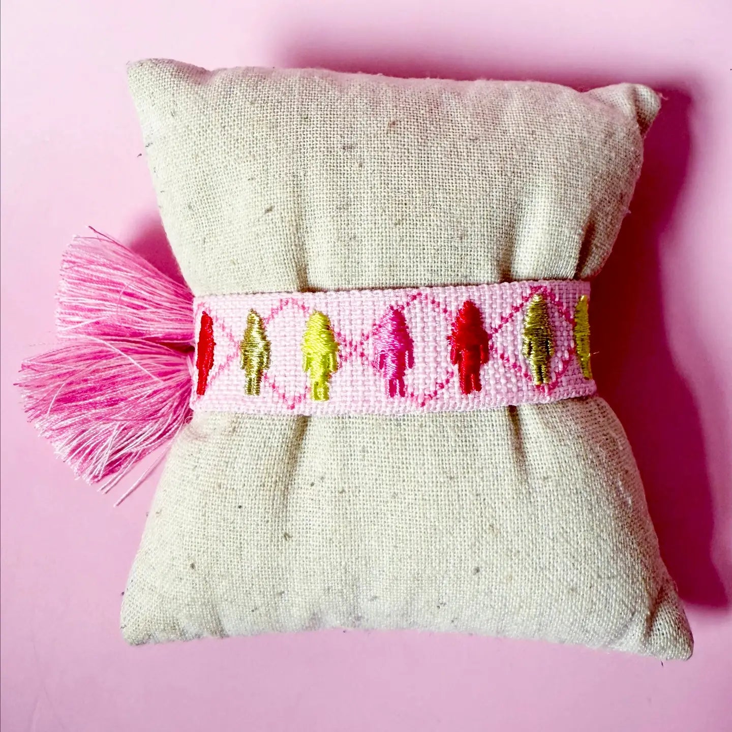 A light pink woven bracelet featuring colorful embroidered nutcracker figures and pink tassels, displayed on a small tan fabric cushion.