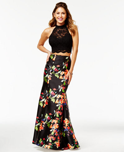 A woman smiling in a two-piece evening gown featuring a black lace halter-neck crop top and a floor-length black satin skirt with a vibrant orange and pink floral print.