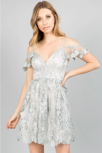 A woman in a short, pale blue cold-shoulder dress with a V-neckline, delicate floral lace overlays, and ruffled sleeves, posing against a light gray background.