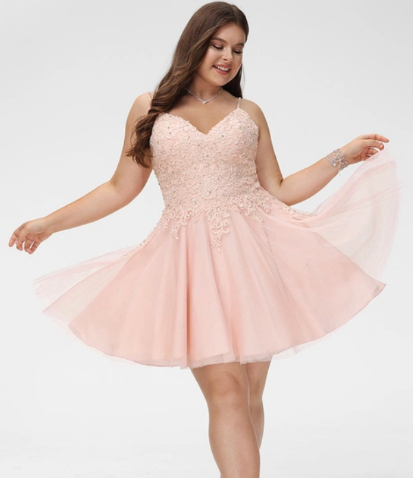 A woman in a short, pale pink cocktail dress with a lace-embellished bodice and a flared tulle skirt, posing against a white background.