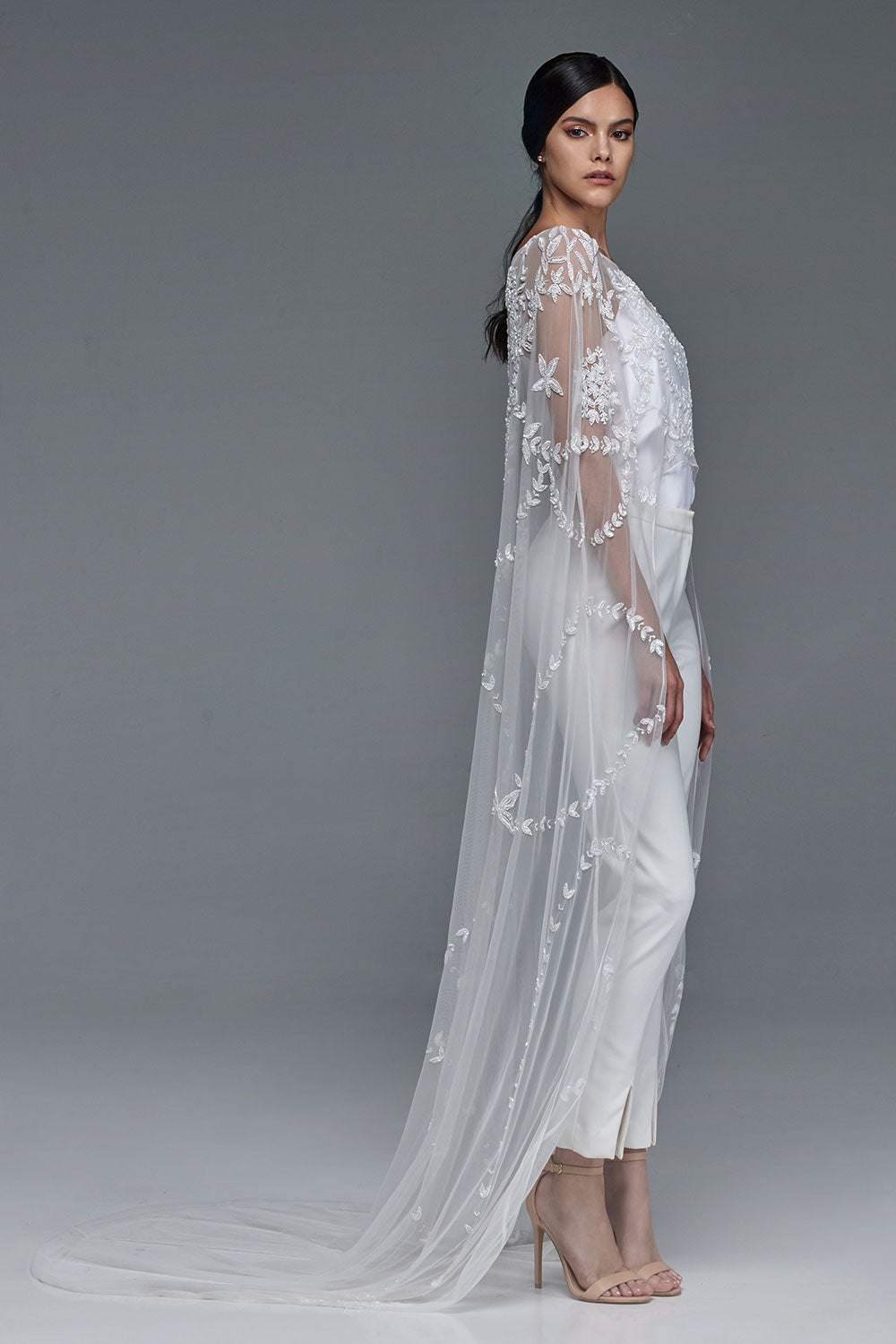 Model in profile wearing a floor-length sheer bridal cape with floral lace embroidery over white tailored trousers.