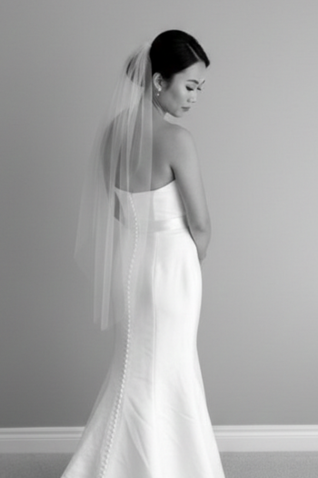 A black and white photo of a bride from the back, wearing a strapless gown with a long row of buttons down the spine and a sheer, waist-length veil tucked into a sleek low bun.