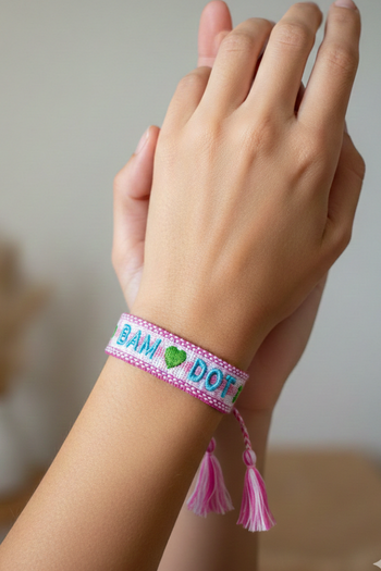 A woven light-pink friendship bracelet featuring "BAM ❤️ DOT" in blue and green embroidery.