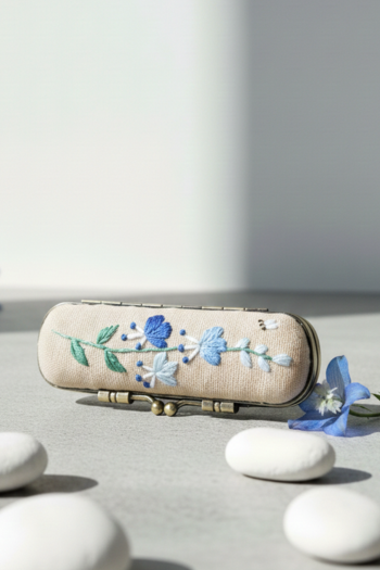 Hand-embroidered lipstick case in beige linen featuring a blue wildflower and a tiny bee. 