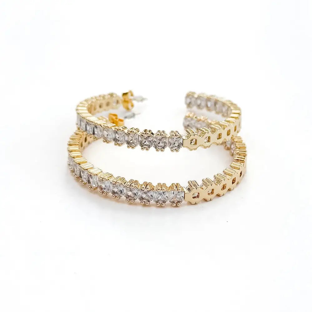 A pair of gold hoop earrings lined with clear, square-cut stones on a white background.