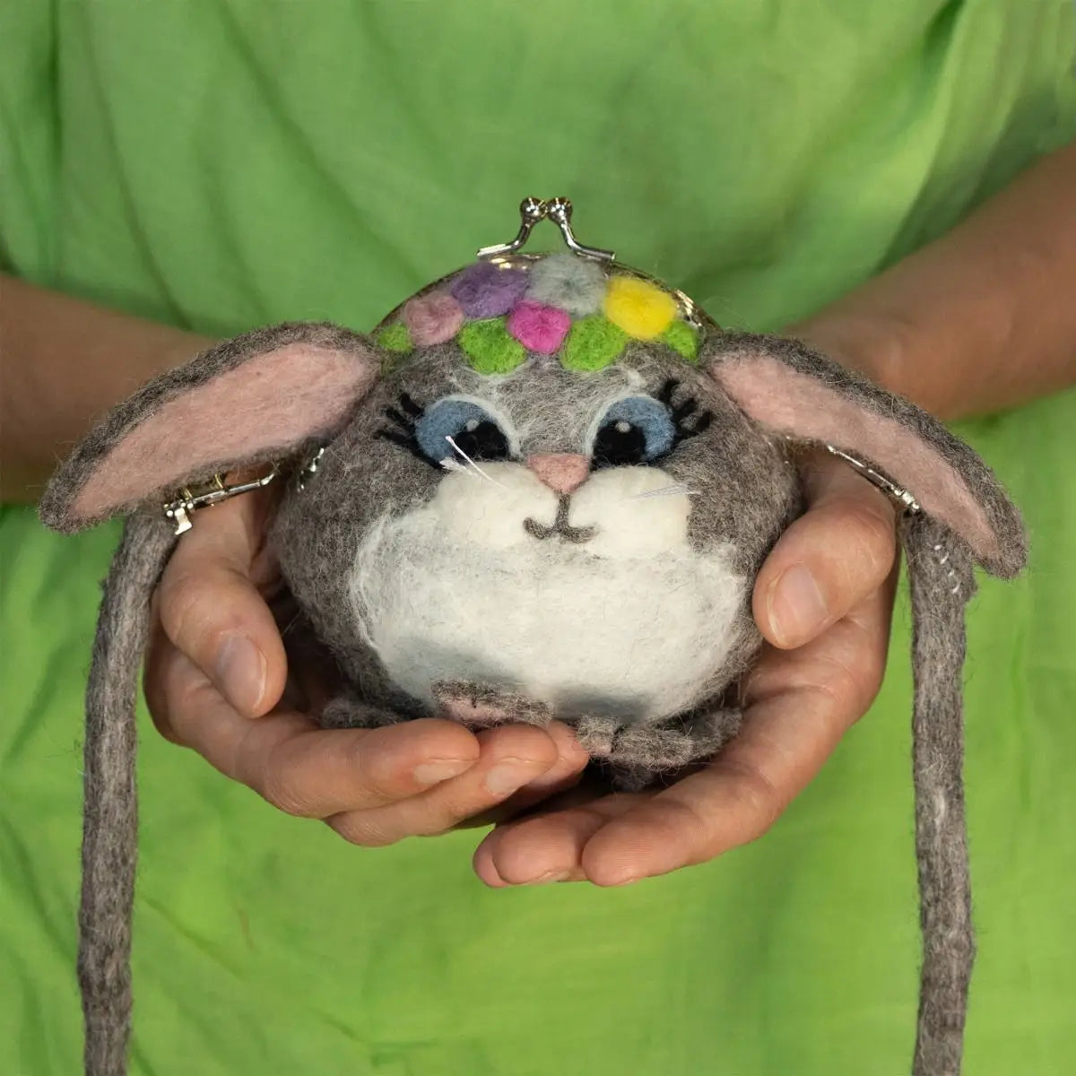A round, grey felt purse shaped like a bunny with a colorful flower crown held in a person's hands.