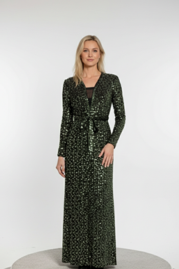 A woman with long blonde hair stands on a white stage, wearing a floor-length, long-sleeved evening gown covered in shimmering dark green and silver sequins. The dress features a deep V-neckline with a black mesh insert and a tied waist detail that creates a flattering, cinched silhouette.