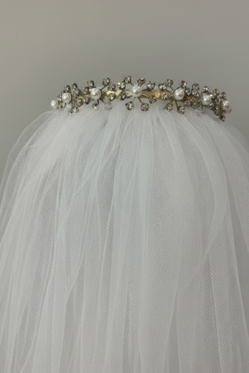 A silver-toned bridal tiara with crystals and pearls, attached to a voluminous white tulle veil, displayed against a minimalist gray background.