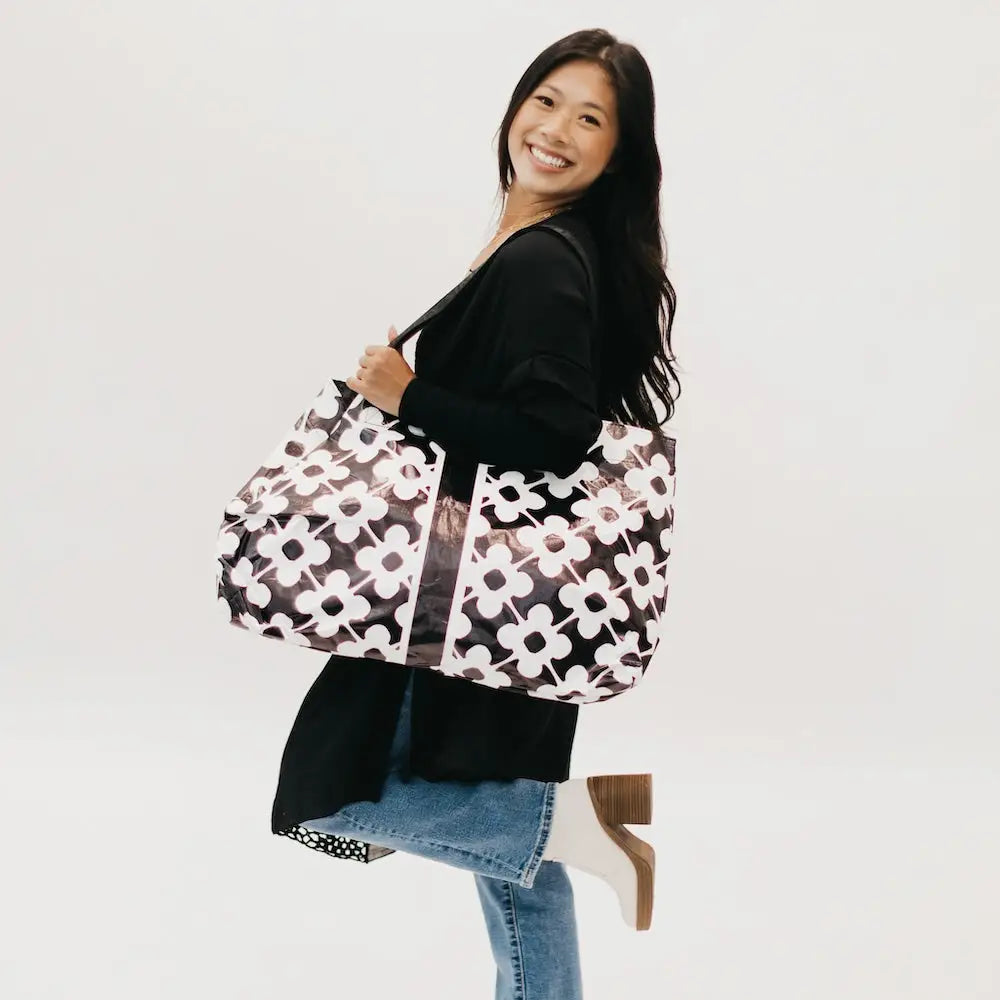 A woman in a black top and jeans carries an oversized black tote bag featuring a bold, white clover floral print and wide shoulder straps.