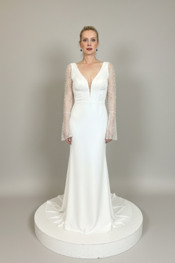 Full-length photo of a woman in a bridal shop wearing a white V-neck wedding dress with long, beaded sleeves and a flowing train.