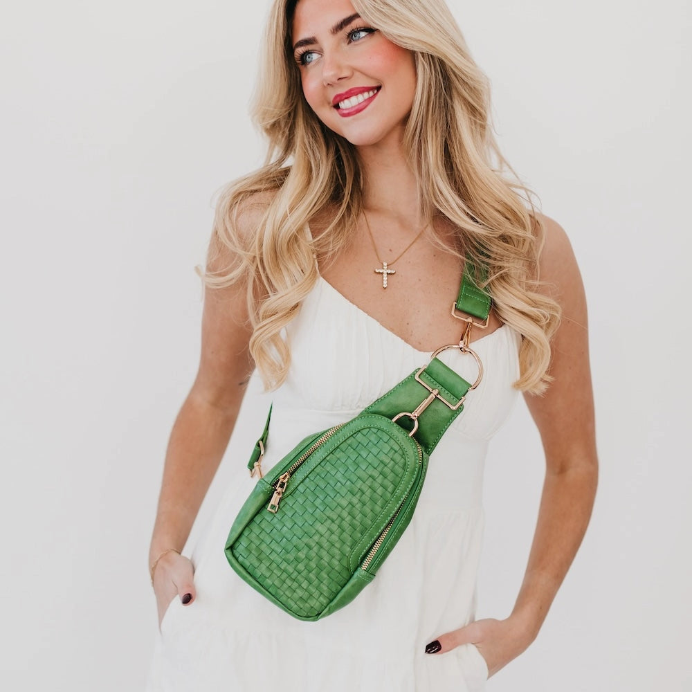 A woman wears an emerald green woven crossbody bag featuring a rounded silhouette, gold-tone zippers, and a wide adjustable strap with a large gold ring detail.