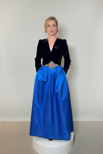 A floor-length gown featuring a long-sleeved black velvet bodice with a V-neckline, an ornate jeweled waist detail, and a voluminous electric blue satin skirt.