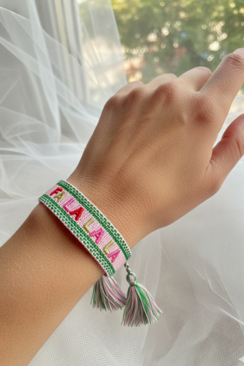 A woven friendship bracelet on a person's wrist featuring "FALALALA" embroidered in red, pink, and gold letters, bordered by green stitched stripes and finished with colorful tassels.