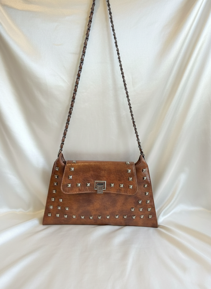 A brown, studded leather handbag with a silver clasp, displayed against a draped, minimalist cream-colored satin background.