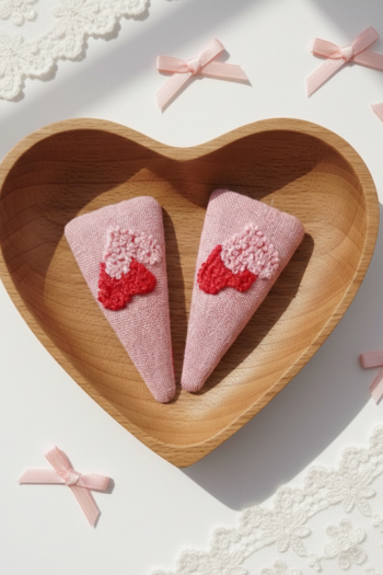 A pair of triangular pink fabric hair clips featuring two hand-embroidered hearts in shades of red and light pink.