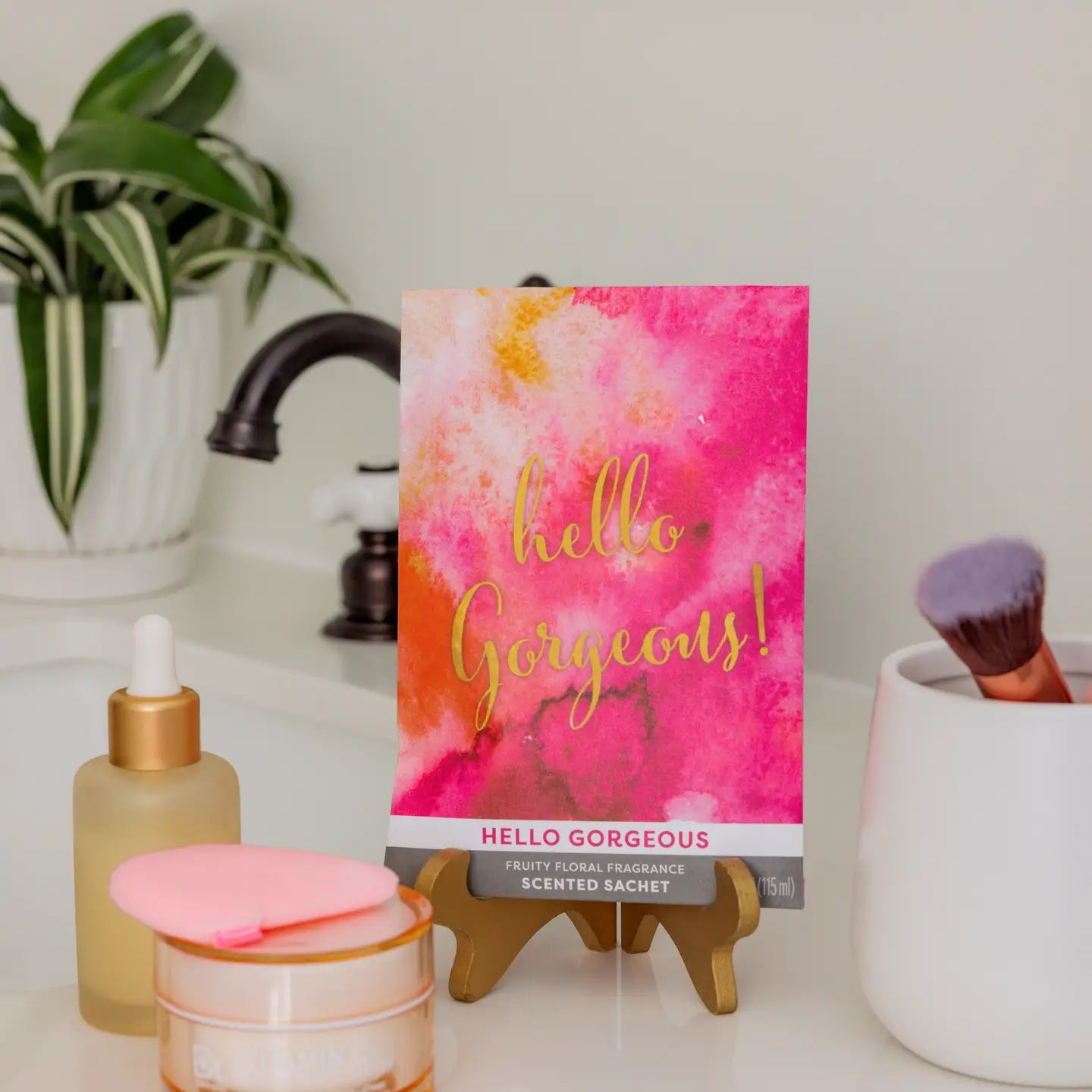 Scented sachet with 'hello gorgeous!' text on a bathroom counter with skincare products and a plant.