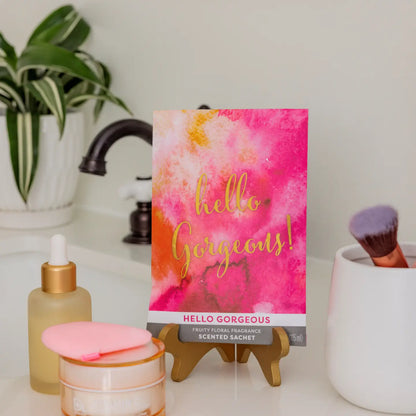 Scented sachet with 'hello gorgeous!' text on a bathroom counter with skincare products and a plant.