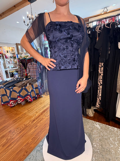 A navy blue evening gown displayed on a mannequin in a clothing boutique. The dress features a floral applique bodice with thin spaghetti straps, a square neckline, and a long, flowing skirt.