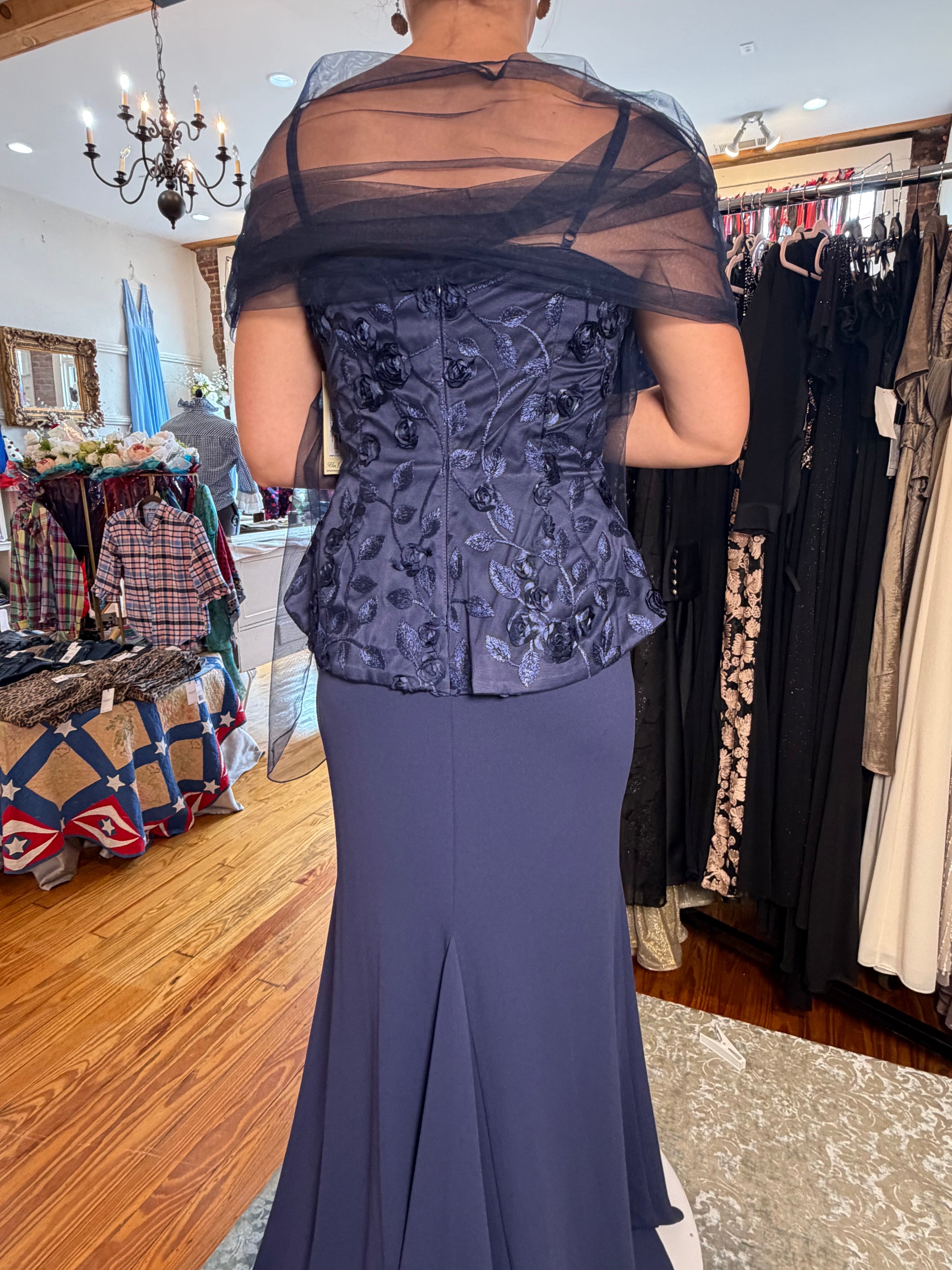 A woman shown from the back wearing a formal evening gown in a clothing boutique. The dress features a dark slate blue floral embroidered corset-style bodice with a sheer black mesh draped shoulder overlay and a fitted mermaid skirt.