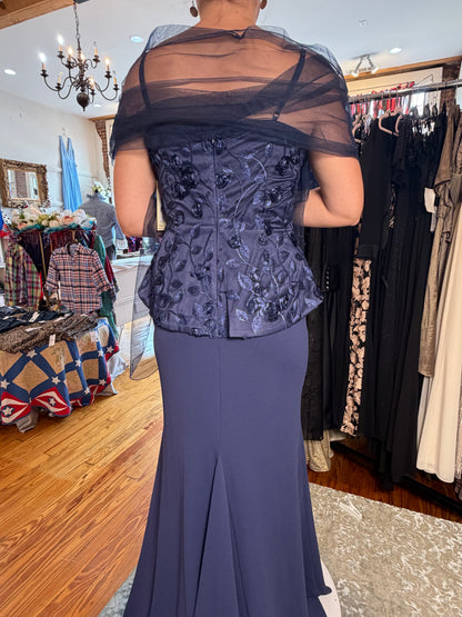 A woman shown from the back wearing a formal evening gown in a clothing boutique. The dress features a dark slate blue floral embroidered corset-style bodice with a sheer black mesh draped shoulder overlay and a fitted mermaid skirt.