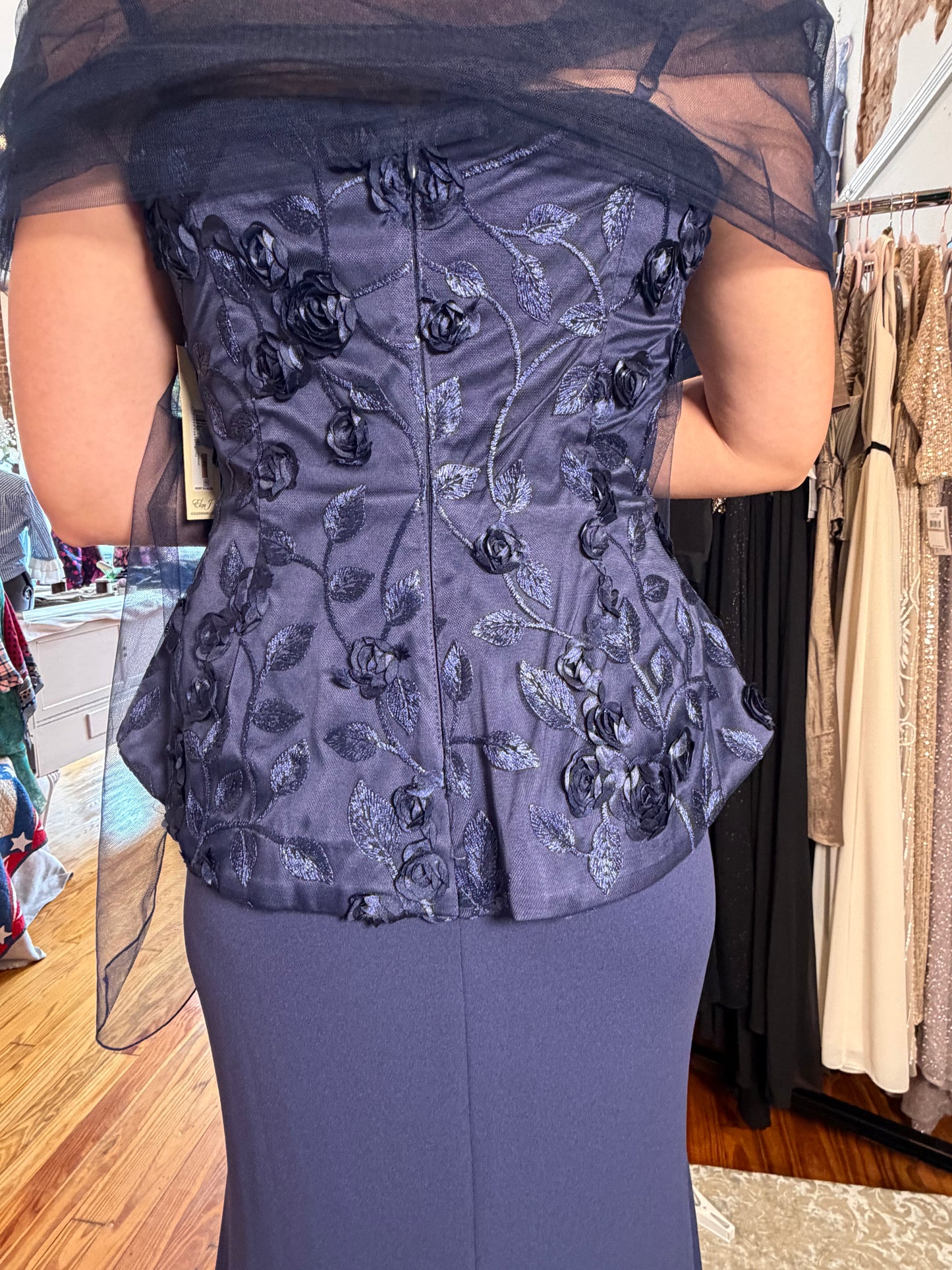 A woman viewed from the back wearing a dark slate blue floral embroidered corset top with sheer black draped sleeves, paired with a matching dark blue skirt in a clothing store.