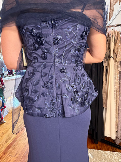 A woman viewed from the back wearing a dark slate blue floral embroidered corset top with sheer black draped sleeves, paired with a matching dark blue skirt in a clothing store.