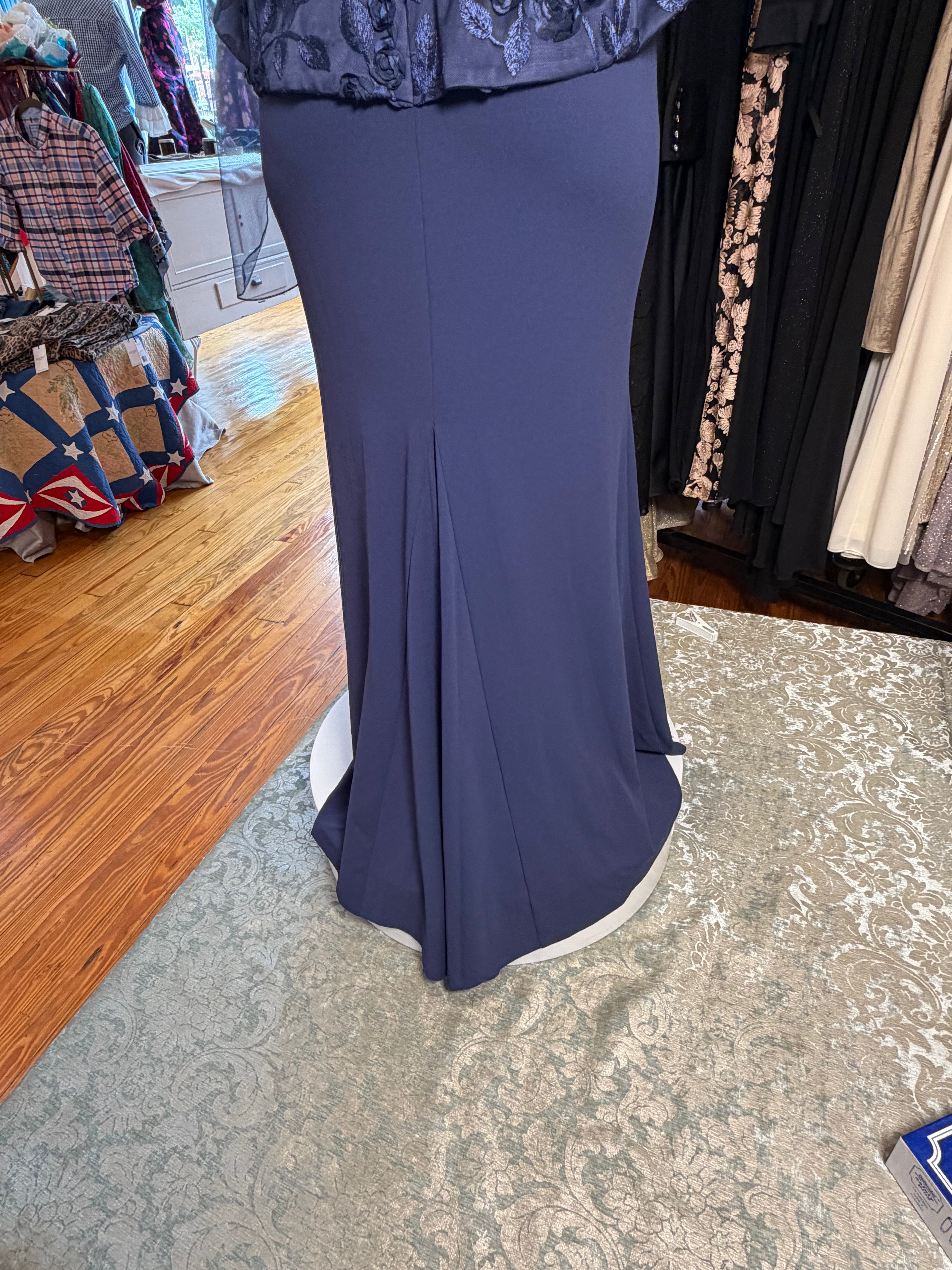 A navy blue floor-length mermaid-style gown displayed on a mannequin in a clothing boutique, with wood floors, patterned rugs, and racks of other garments visible in the background.