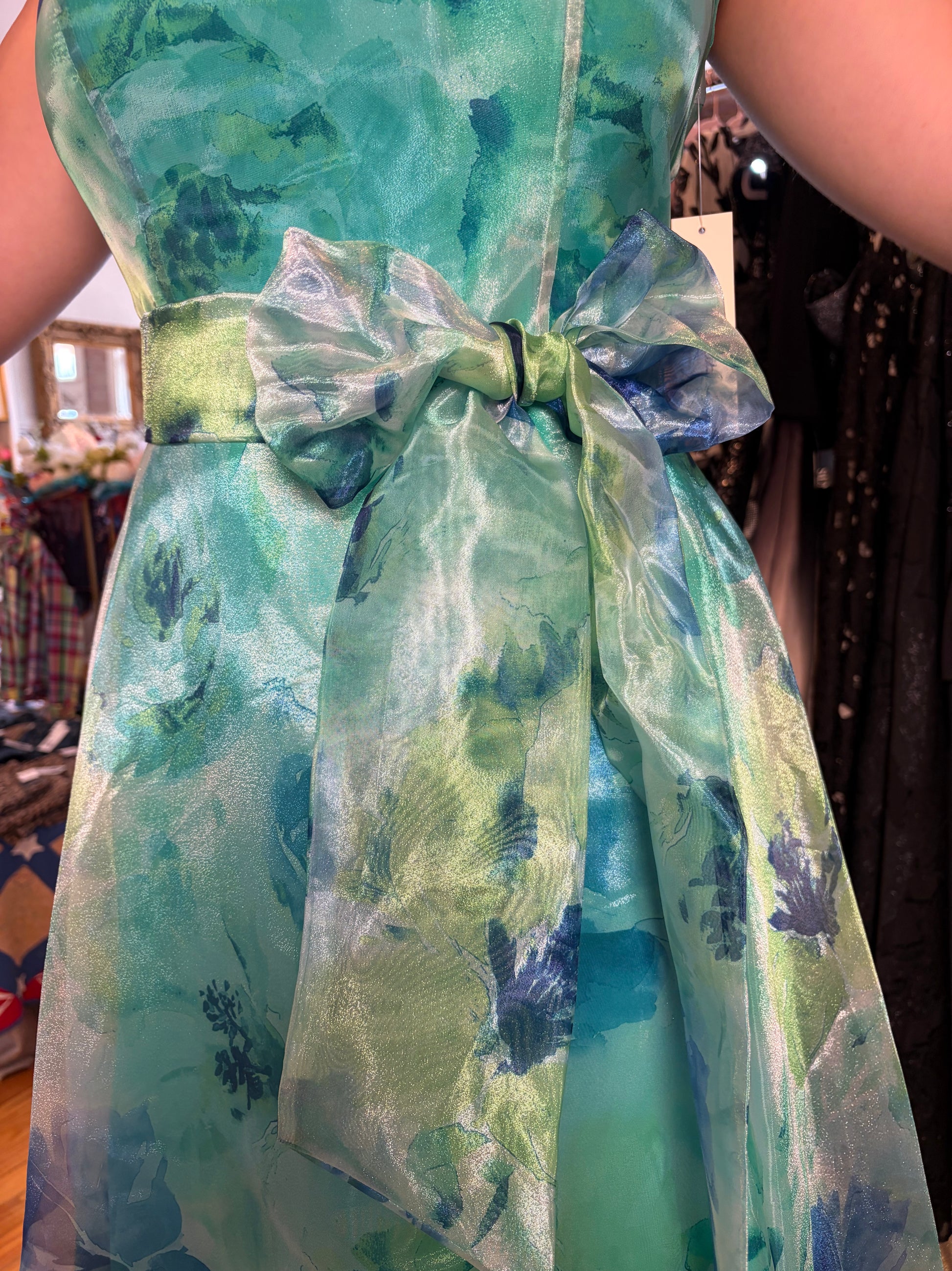 A close-up of a sleeveless dress with a watercolor-style green and blue pattern, featuring a wide waistband and a large bow at the center front.