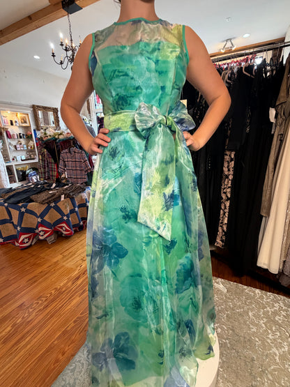 A sleeveless green watercolor floral dress with a sheer mesh bodice and a matching waist bow, modeled in a clothing boutique.