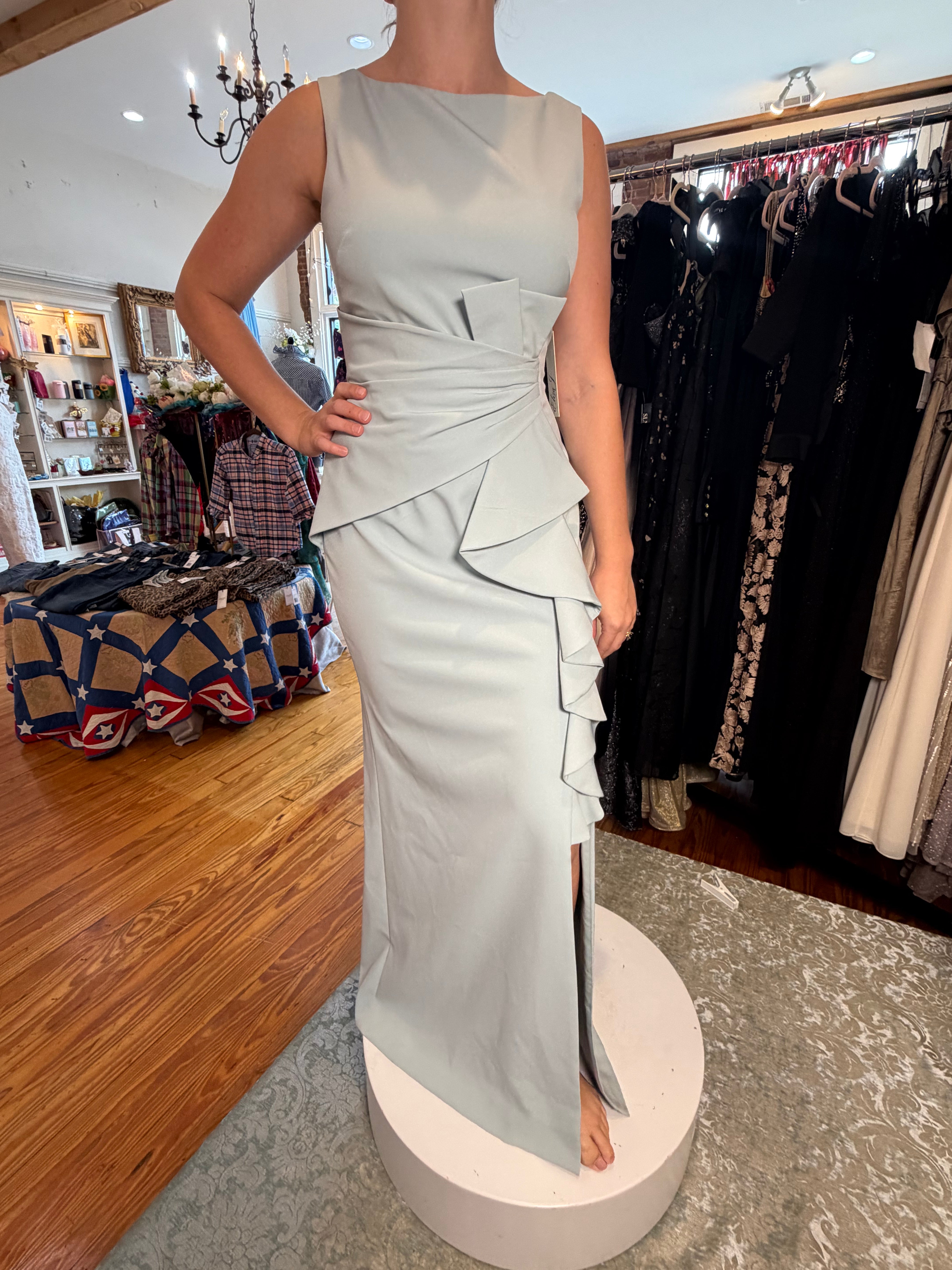 A sleeveless light sage green evening gown with a draped ruffle waist and side slit, displayed on a white pedestal inside a clothing boutique.