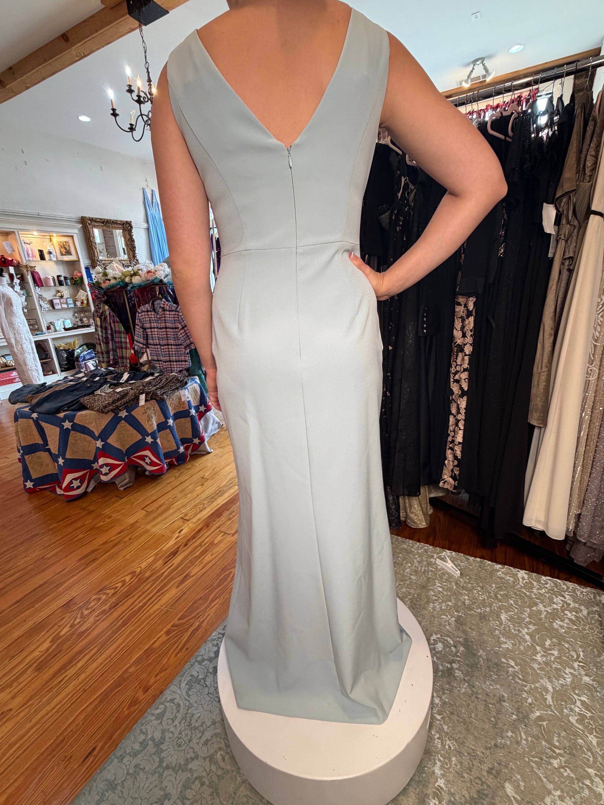 A sleeveless, floor-length light sage green gown with a deep V-neckline, shown from the back on a white circular pedestal inside a clothing boutique.