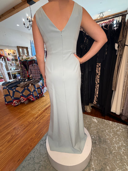 A sleeveless, floor-length light sage green gown with a deep V-neckline, shown from the back on a white circular pedestal inside a clothing boutique.