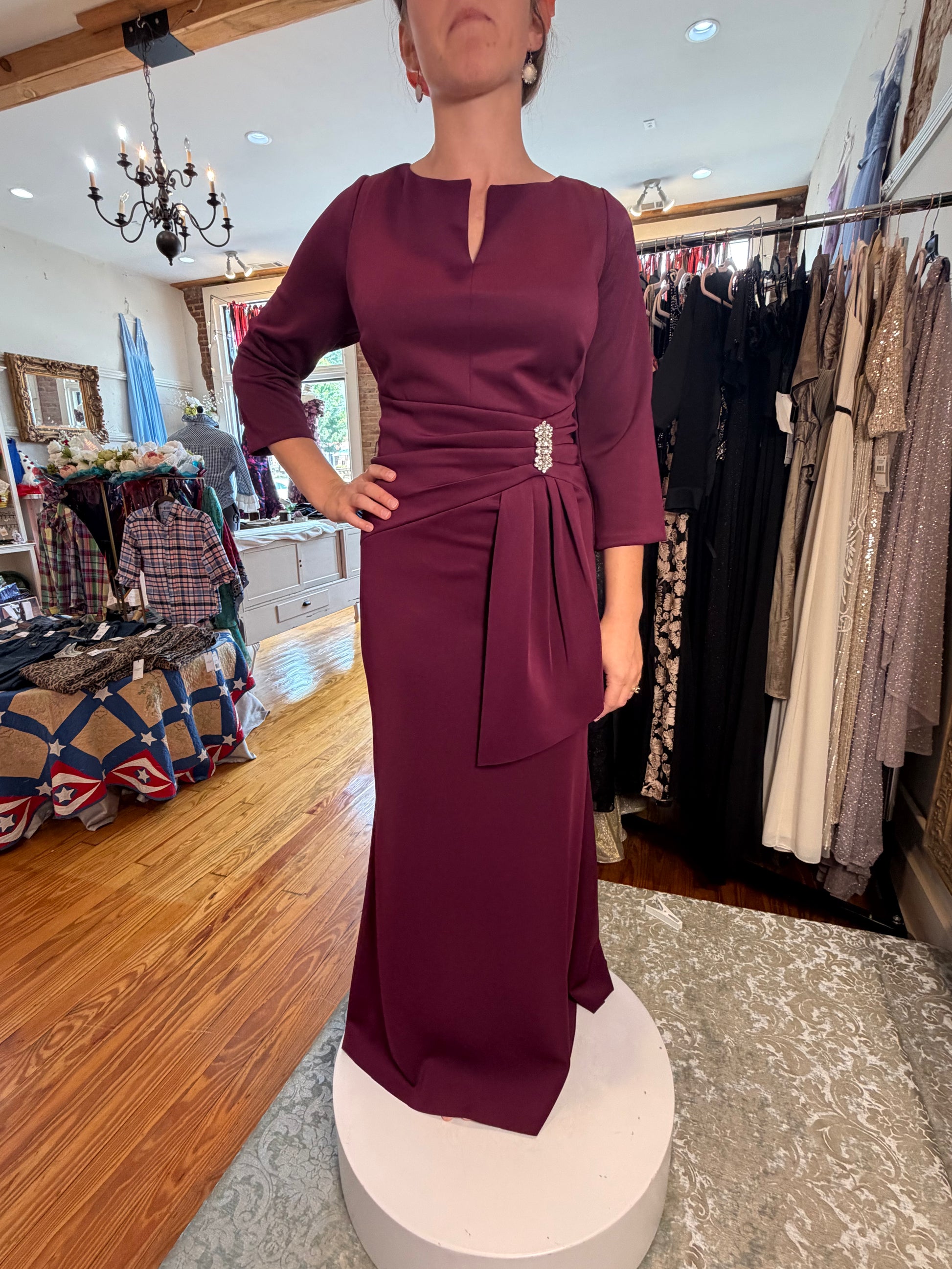 A woman wearing a floor-length burgundy dress with three-quarter sleeves, a split neckline, and a self-tie waist belt with a jeweled embellishment, standing on a white pedestal inside a clothing boutique.