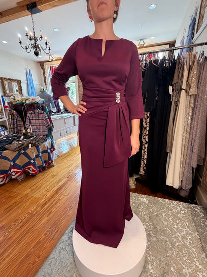 A woman wearing a floor-length burgundy dress with three-quarter sleeves, a split neckline, and a self-tie waist belt with a jeweled embellishment, standing on a white pedestal inside a clothing boutique.