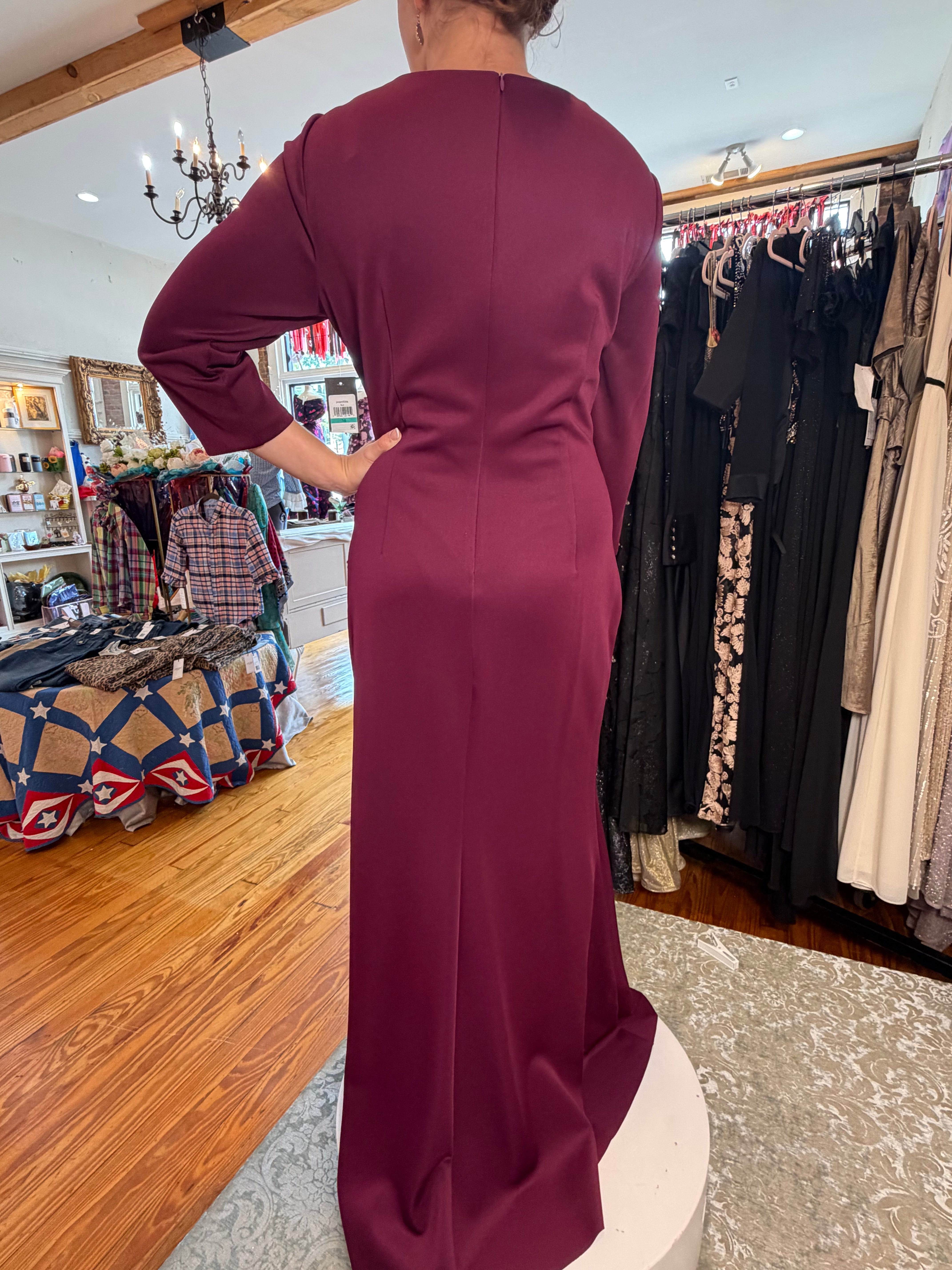 A woman wearing a long-sleeve, floor-length burgundy dress, photographed from behind inside a clothing boutique. The dress has a straight, fitted silhouette with a subtle sheen and a single seam down the back.