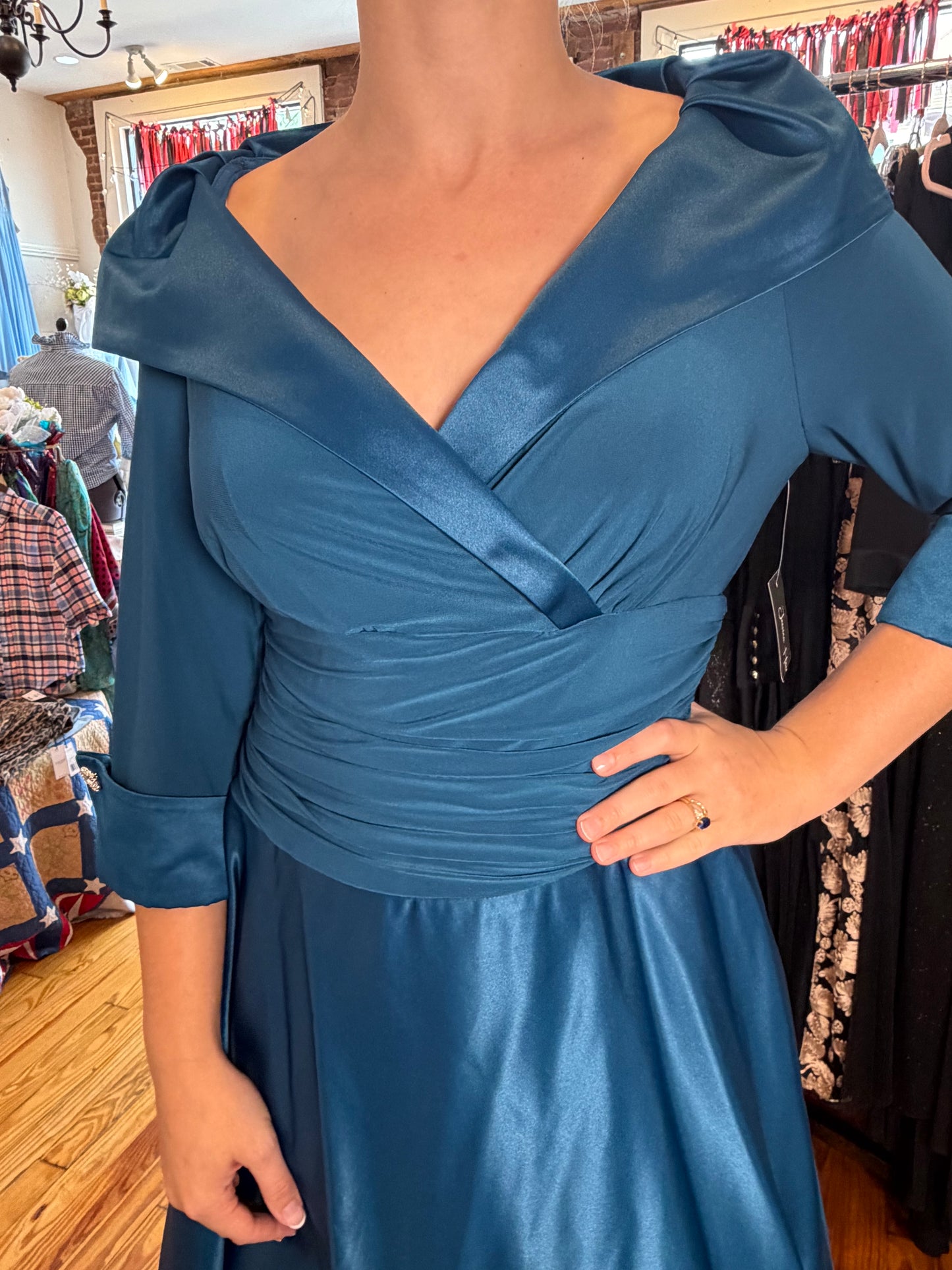 A woman wearing a teal blue satin dress with a wrap-style V-neck bodice, puffed shoulder details, and gathered waist, photographed inside a clothing boutique.