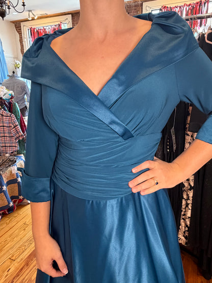 A woman wearing a teal blue satin dress with a wrap-style V-neck bodice, puffed shoulder details, and gathered waist, photographed inside a clothing boutique.