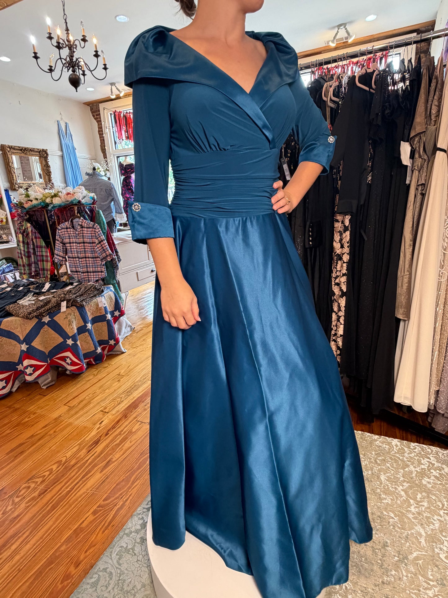 A woman wearing a floor-length teal blue gown with a wrap-style bodice, puffed shoulders, and a full skirt, standing inside a clothing boutique with racks of dresses in the background.