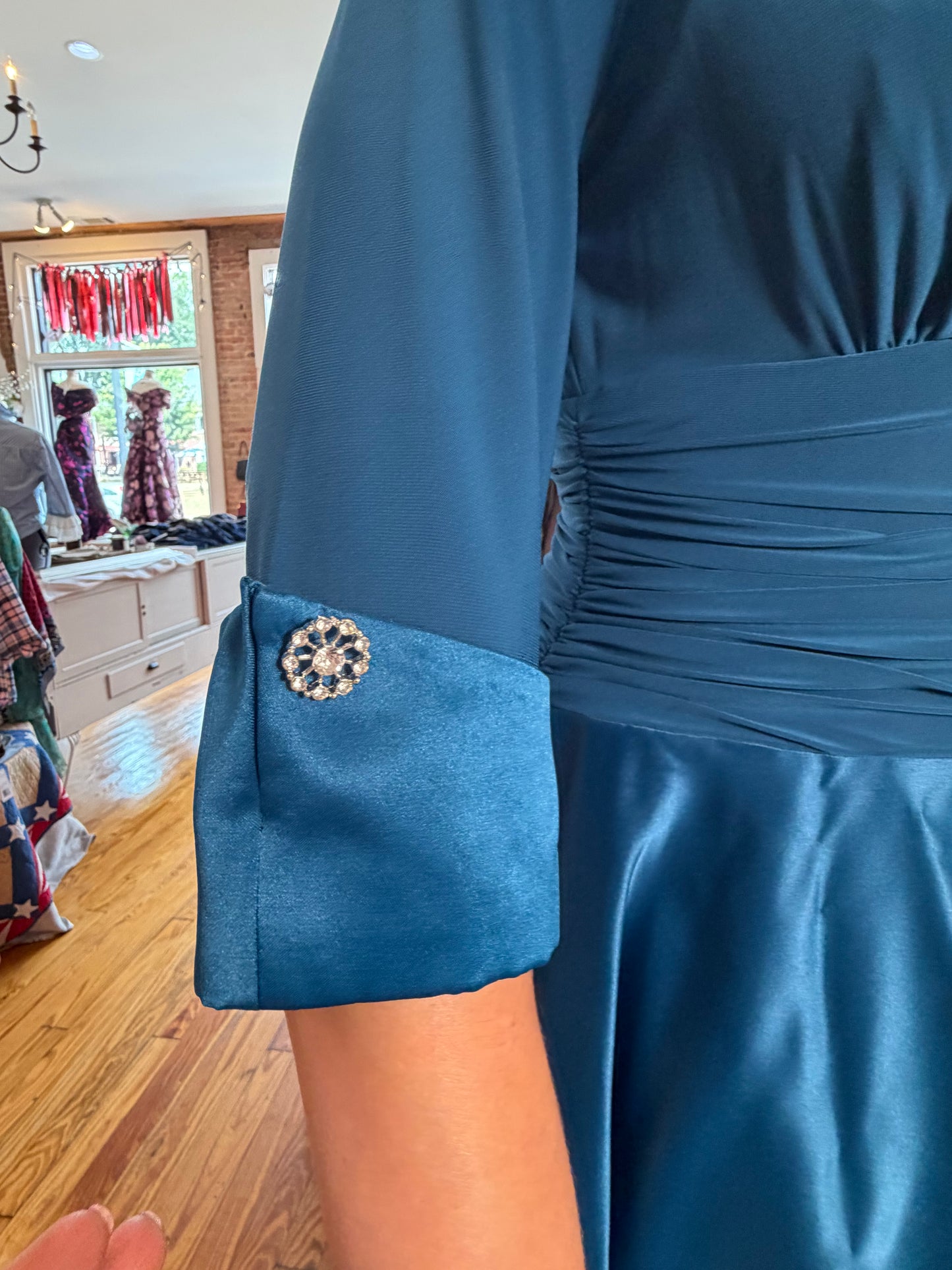 A close-up of a person wearing a teal blue dress with a gathered waist and a rolled cuff featuring a decorative jeweled brooch, in a boutique setting.