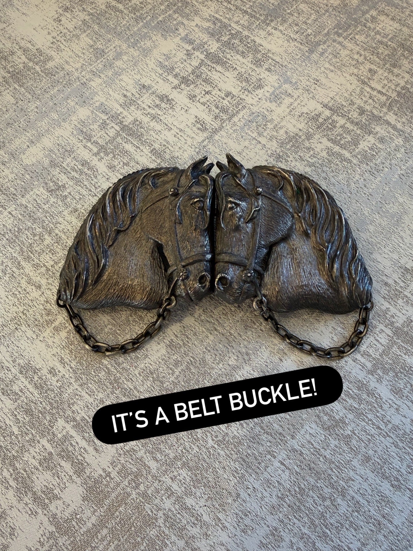 silver-toned metal belt buckle shaped like two horse heads facing each other in profile and connected by a chain. A black text box at the bottom reads 