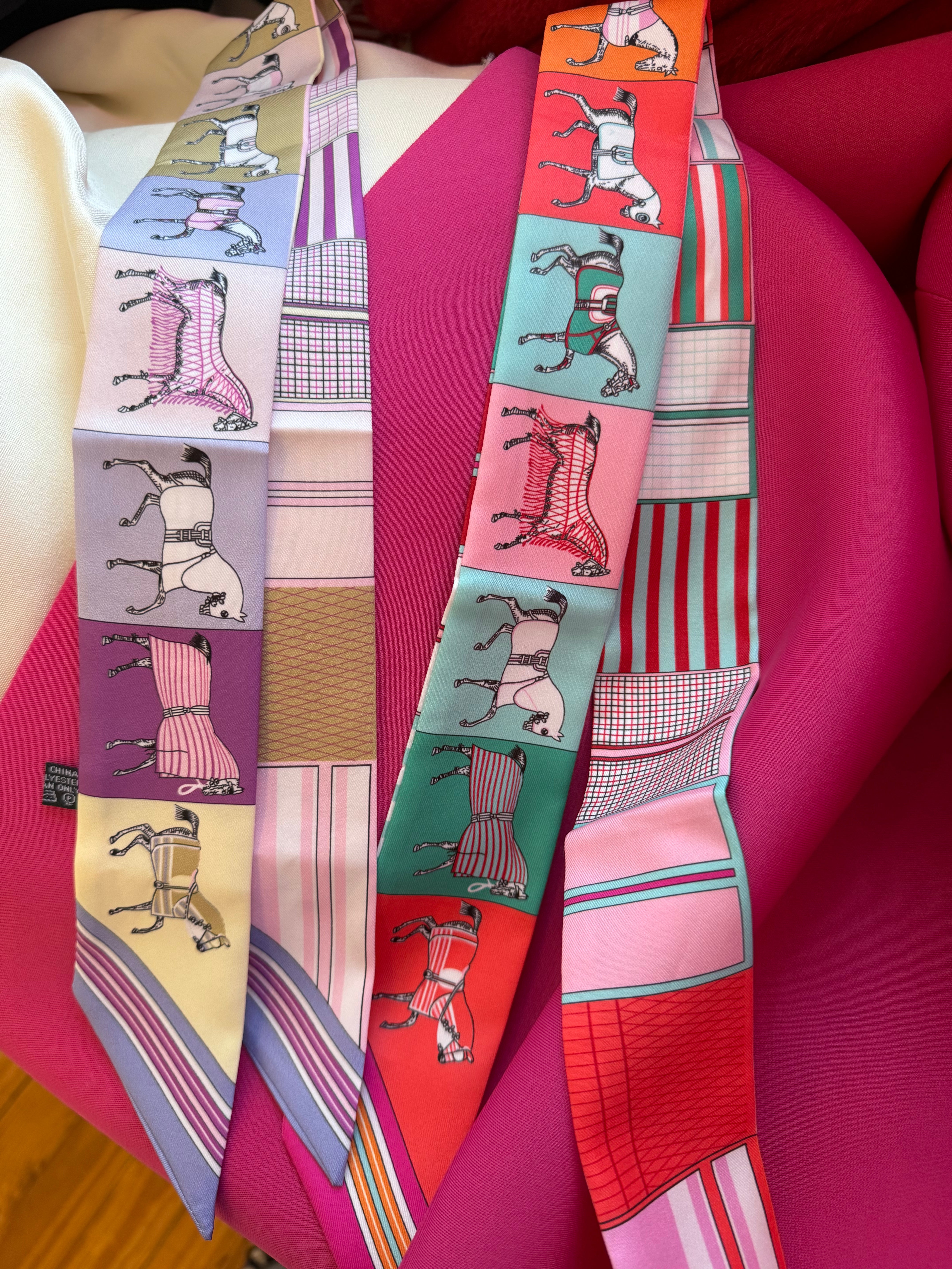 Two narrow silk scarves featuring horse illustrations and geometric patterns draped on a bright pink background.