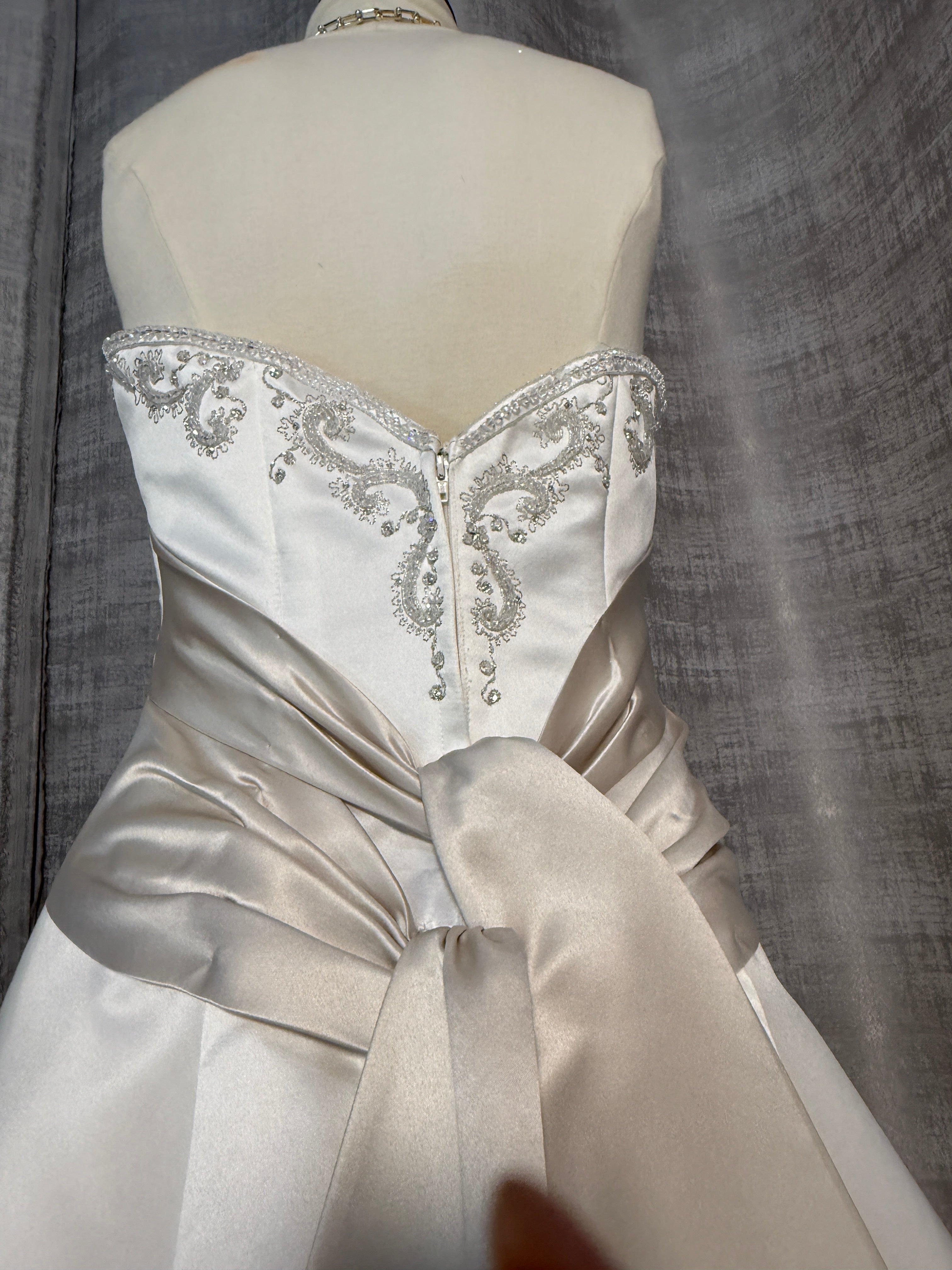 Close-up of Mon Cheri wedding gown bodice showing intricate silver beaded and embroidered scrollwork on ivory satin sweetheart neckline, center-front zipper, and champagne ruched sash with bow knot detail
