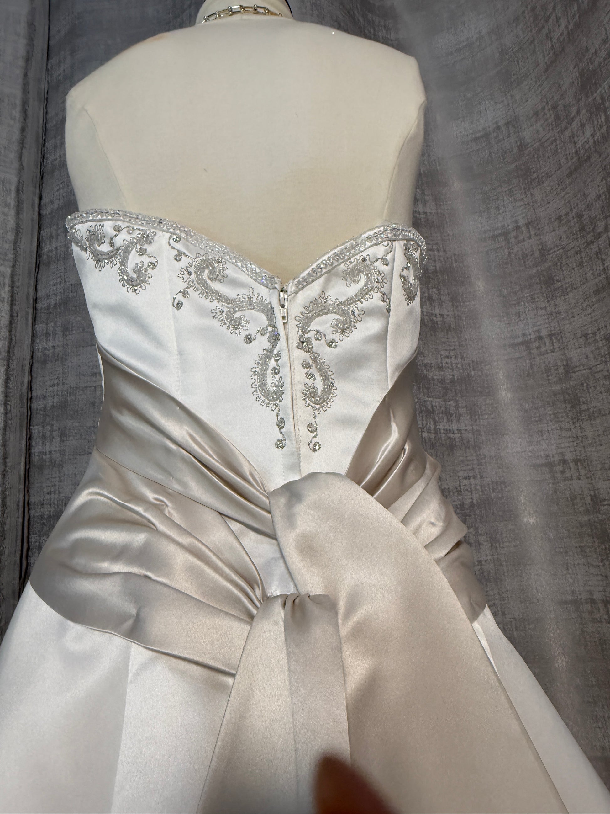 Close-up of Mon Cheri wedding gown bodice showing intricate silver beaded and embroidered scrollwork on ivory satin sweetheart neckline, center-front zipper, and champagne ruched sash with bow knot detail