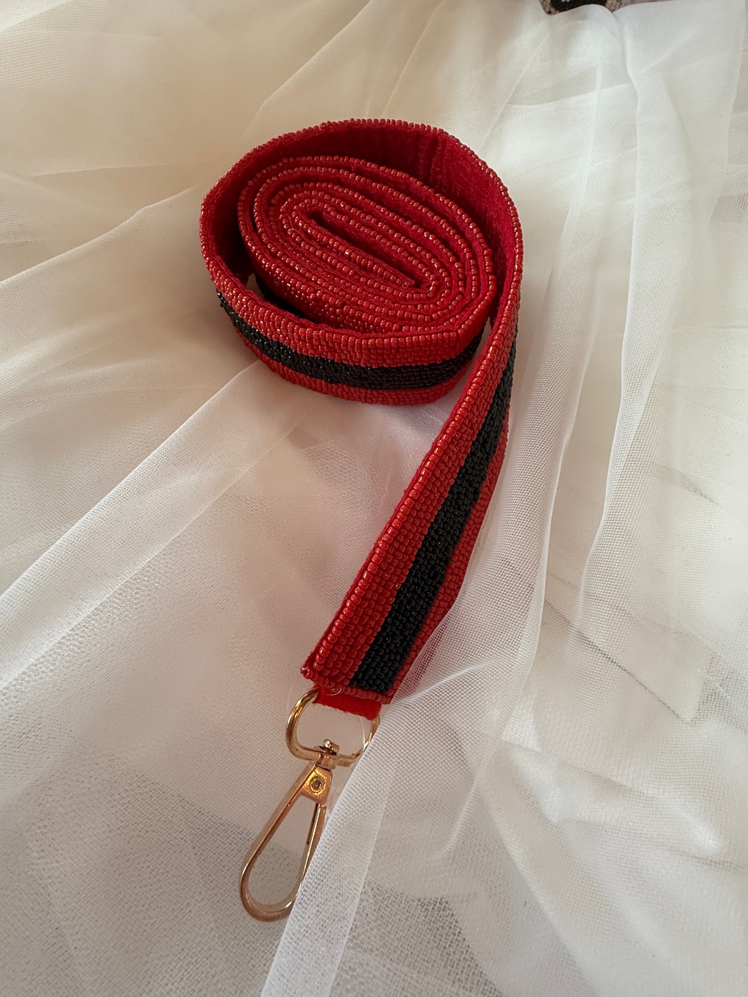 A close-up of a thick, red beaded strap featuring a solid black beaded stripe running down the center, finished with a gold-tone lobster clasp and displayed against a white mesh background.