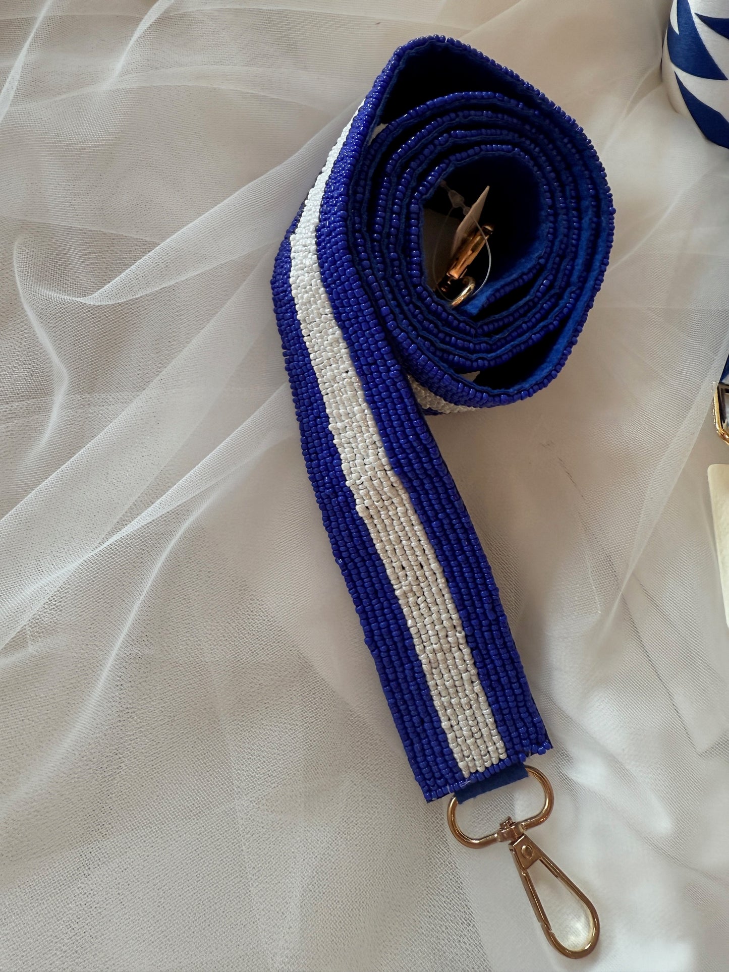 A thick, royal blue beaded strap featuring a solid white beaded stripe running down the center, finished with a gold-tone lobster clasp and coiled against a white mesh background.