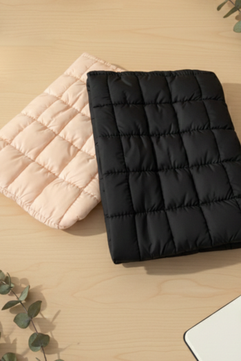 Two rectangular, quilted puffer pouches—one in matte black and one in light peach—resting diagonally on a light wood surface. The pouches feature a distinct square grid-like stitched pattern, creating a soft, padded texture.
