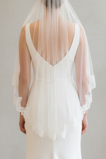 Back view of a bride wearing a classic fingertip-length wedding veil made of sheer white tulle. The veil features a delicate scalloped lace trim along the edges and is gathered at the top