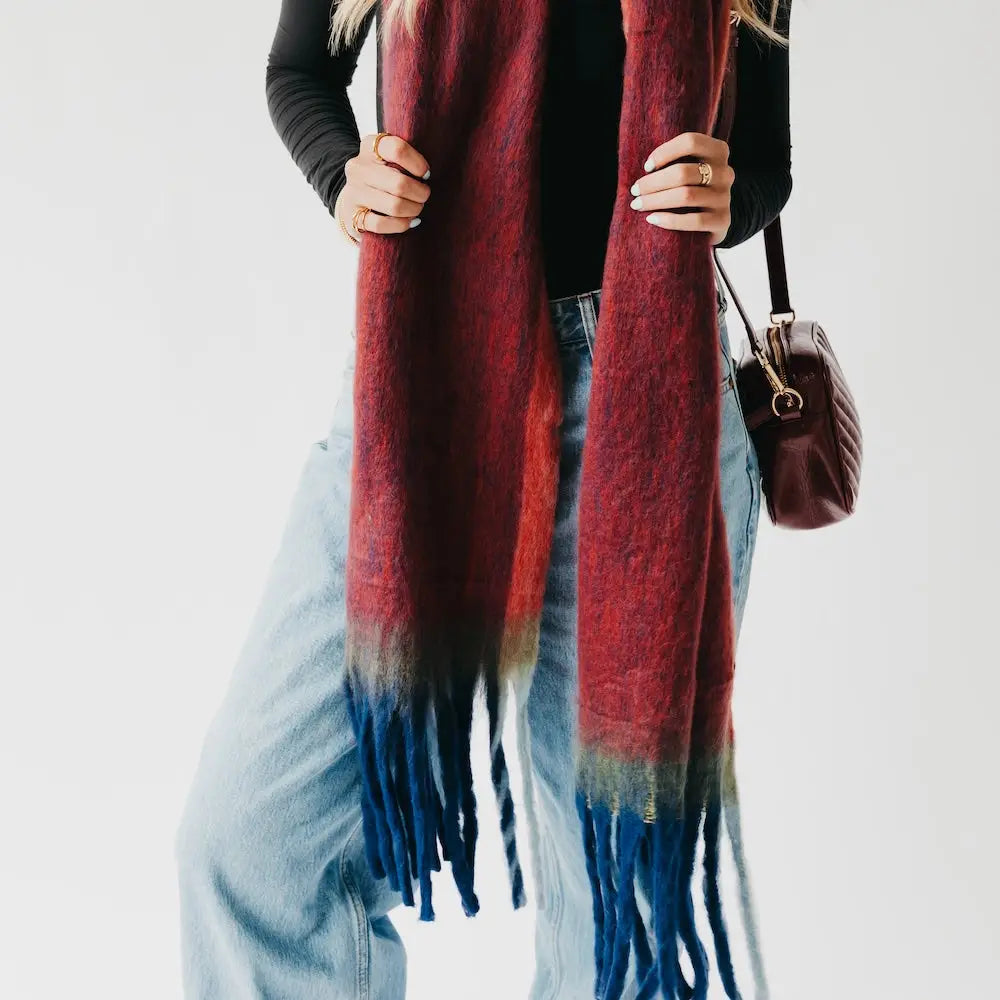 A woman in a black long-sleeved top and light-wash jeans wears an oversized, deep red and navy blue ombre scarf with thick, chunky blue tassels at the ends.